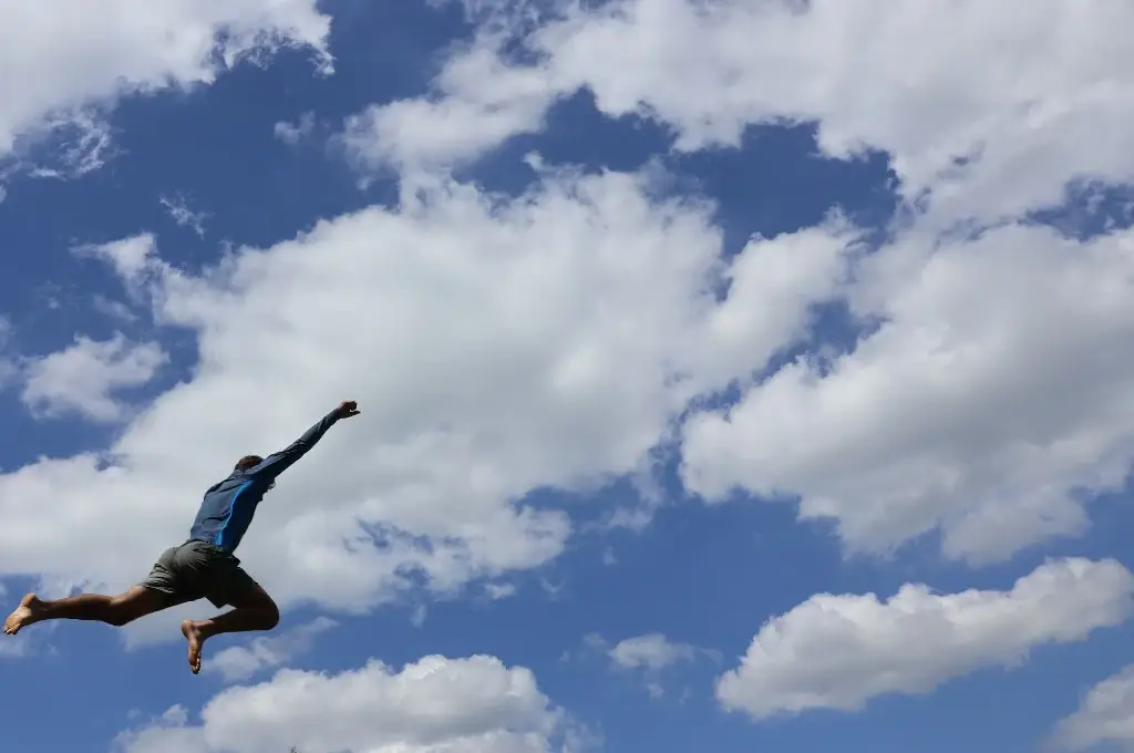 Person wearing a blue top and gray shorts jumps mid-air with arms raised against a blue, cloudy sky.