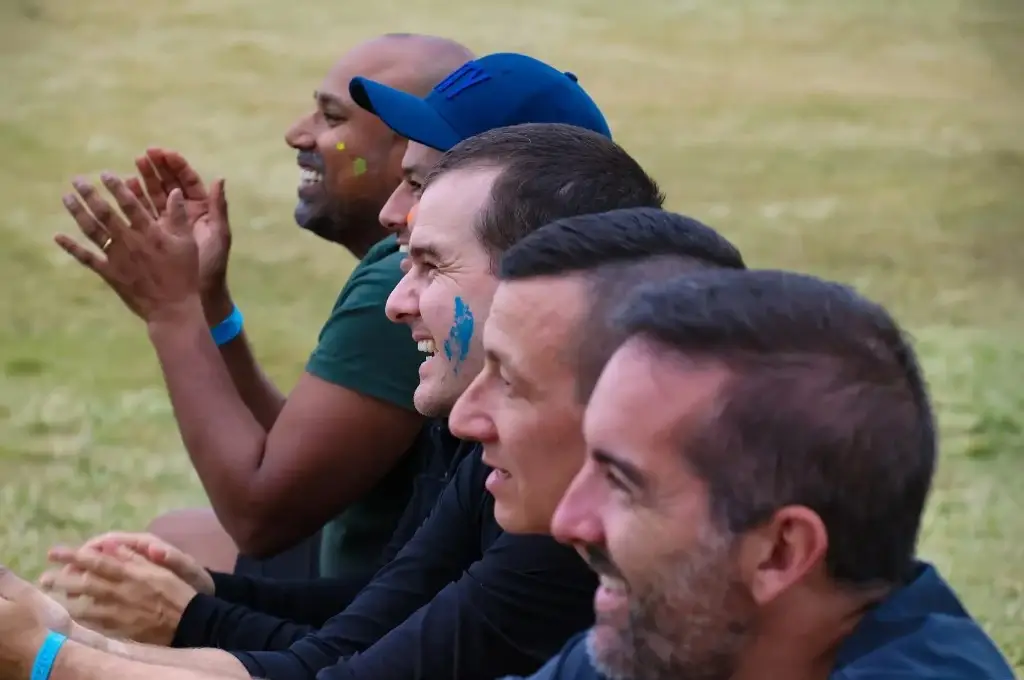 Row of men sitting on grass, clapping and smiling at an outdoor event, one with blue face paint on his cheek.