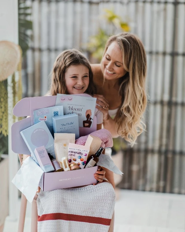 Mother and daughter smile while holding a lavender gift box filled with skincare and books outdoors.