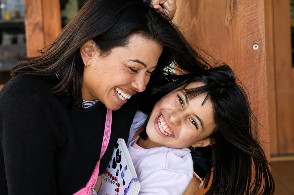 Two women share a joyful hug outside, smiling and leaning together with one holding a small notebook or crafts utensil case between them.