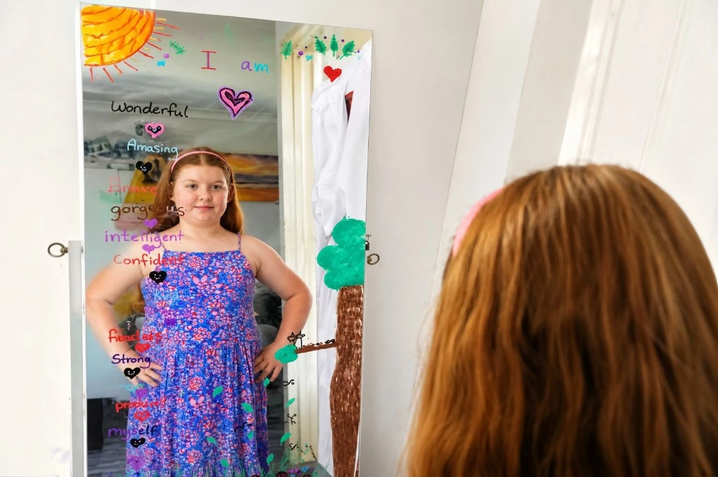 Girl in a purple floral dress stands before a mirror covered with colorful, handwritten affirmations on the glass.