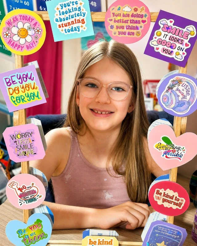 Smiling girl with glasses sits at a table, framed by colorful sticky notes with cheerful messages like 'Be kind' and 'You are amazing.'