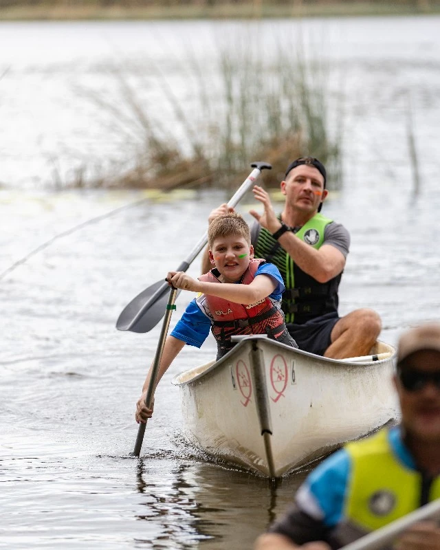 Young boy at the bow of a white canoe paddling on a lake, wearing a life jacket, assisted by an adult behind him.