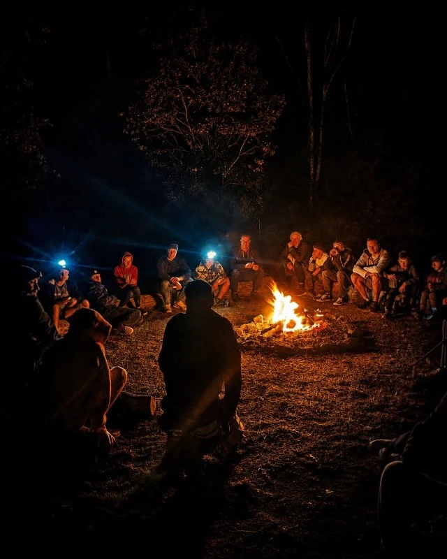 People sit in a circle around a glowing campfire at night in a wooded clearing.