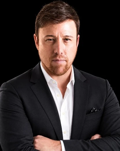 Professional portrait of a man in a dark suit with a white shirt, arms crossed, neutral expression, against a black background of the photo's studio setting