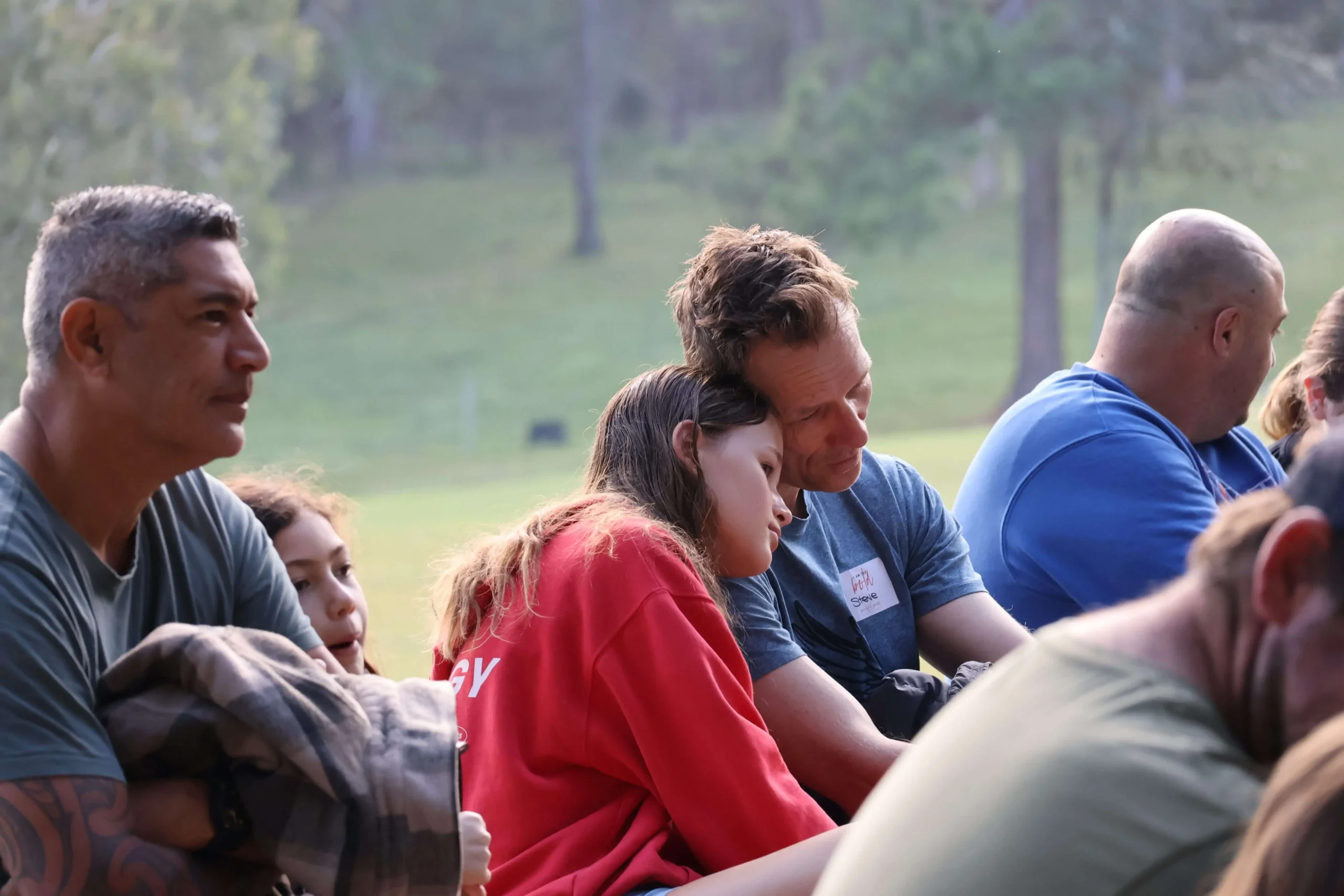Group of people seated outdoors, facing left, talking and listening at what looks like a casual gathering or event.